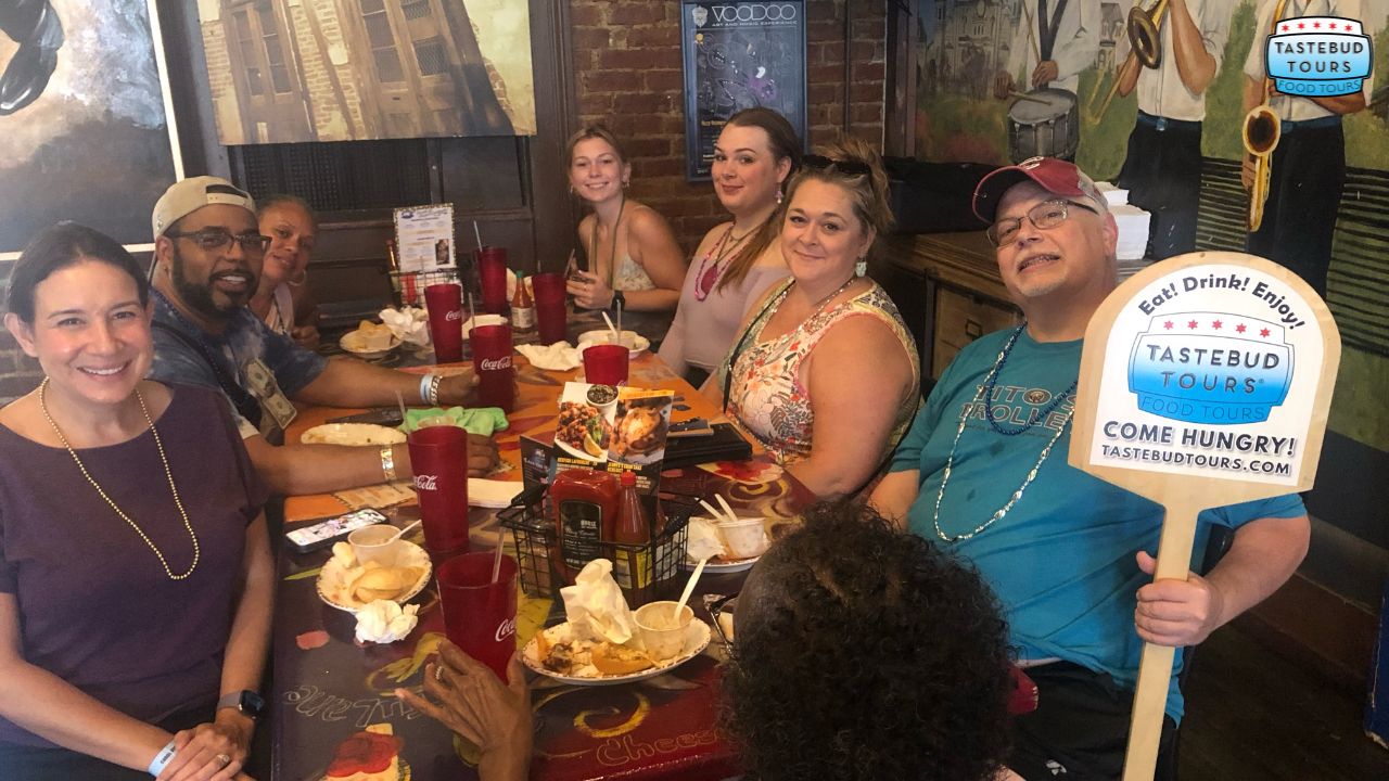 best-time-for-brunch-in-new-orleans (1) Group of people at a restaurant table holding Tastebud Tours sign, smiling and surrounded by food and drinks.