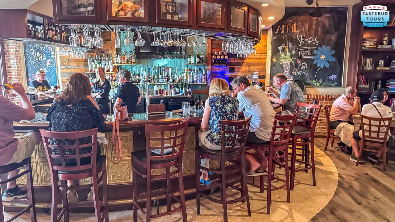 People sitting at a bar with glasses and bottles, chalkboard in background, cozy atmosphere.