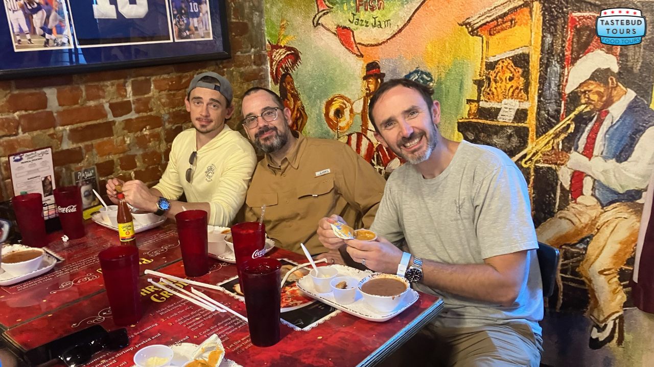 Three men smile at a restaurant table with food and drinks against a colorful mural wall.