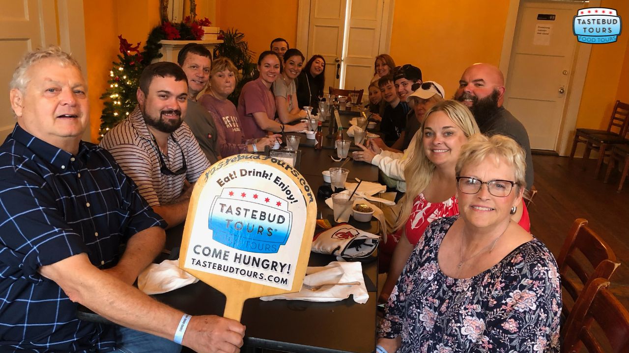 best-time-to-visit-new-orleans-for-food-tour (2) Group of people sitting at a long table holding a Tastebud Tours sign.