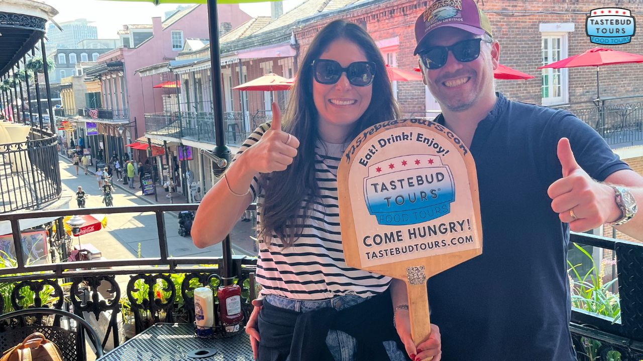 famous-breakfast-restaurant-in-new-orleans (1) Two people holding thumbs up sign for Tastebud Tours on a balcony overlooking a street.