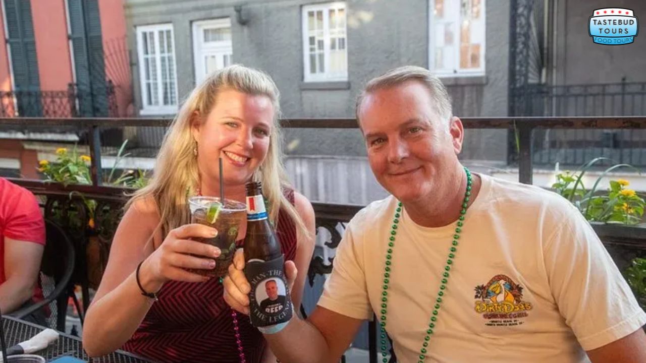 Two people enjoying drinks on a balcony, wearing beads and smiling at the camera.