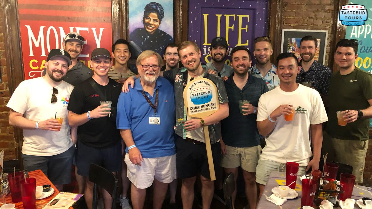 things-to-do-in-new-orleans-this-weekend Group of people posing indoors with drinks, smiling next to colorful posters on a brick wall.