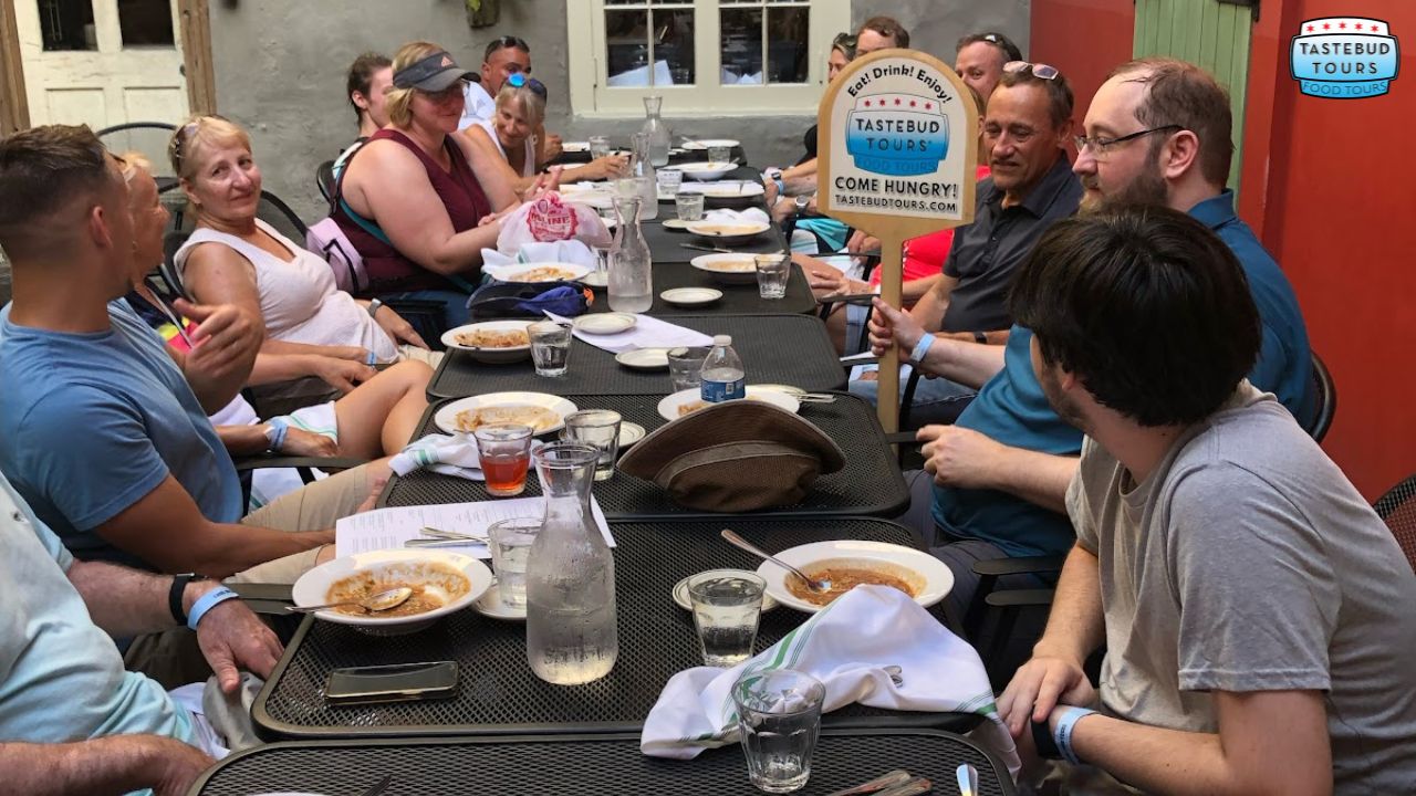 Group at outdoor table enjoying a Tastebud Tours meal, some holding a sign, empty bowls in front.