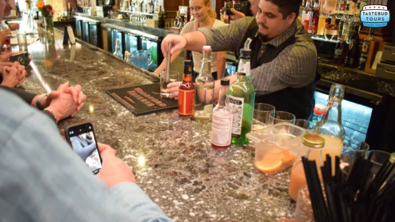 Bartender mixing drinks at a bar, with patrons watching and one taking a photo.