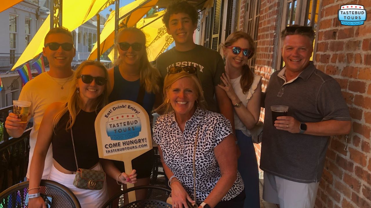 Group of people smiling on a sunny balcony, promoting Tastebud Tours with a sign.