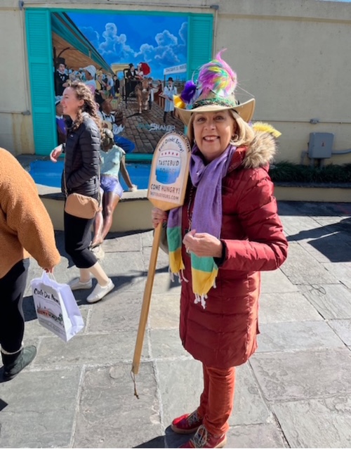 Woman in red coat and colorful hat holds a tour sign, standing on a street with people and a mural in the background.