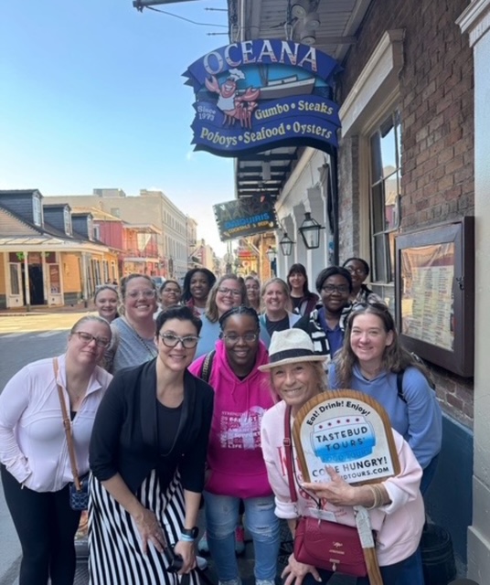 Group of people posing outside Oceana restaurant holding a Tastebud Tours sign.