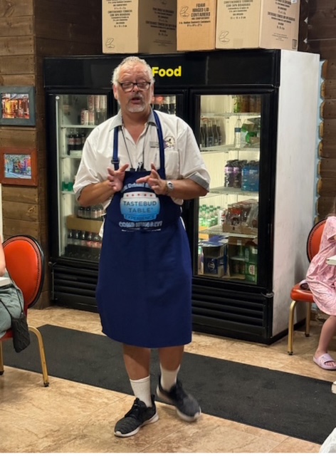 Man with apron speaking in front of a refrigerator in a store setting.