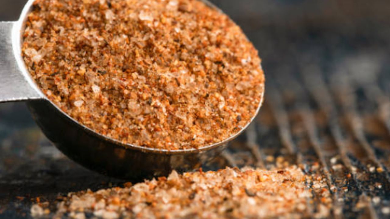 Close-up of spice mix in a metal measuring spoon on a dark surface.