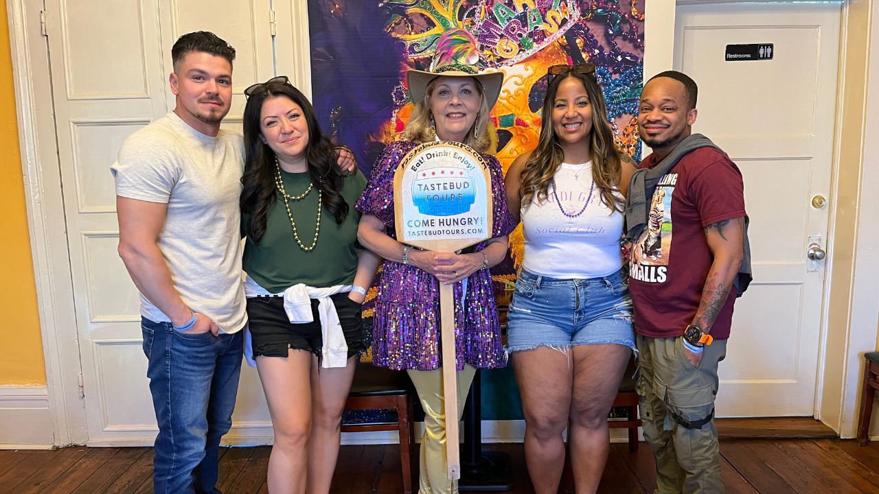Five people stand together indoors, with one holding a sign saying 'Tastebud Tours' and a colorful backdrop.