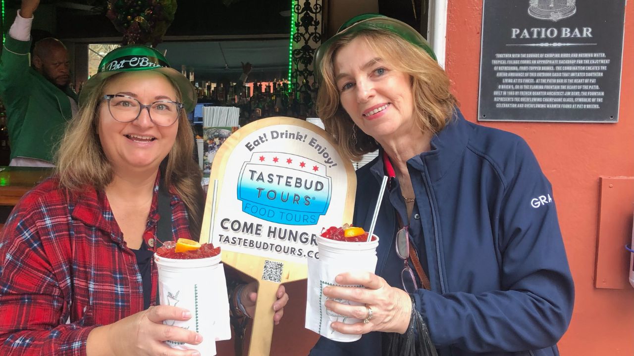Two women holding drinks, smiling with a Tastebud Tours sign between them.