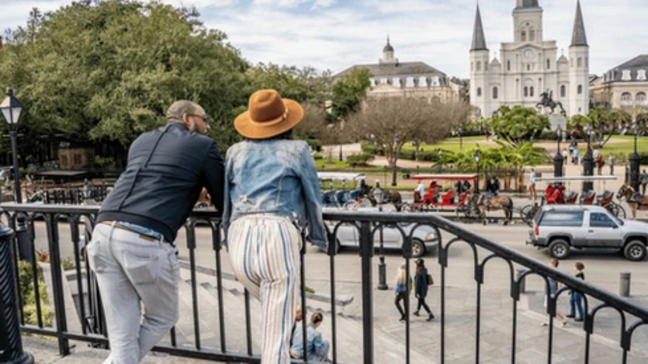 Two people leaning on a railing overlook a city square with a cathedral and horse-drawn carriages.