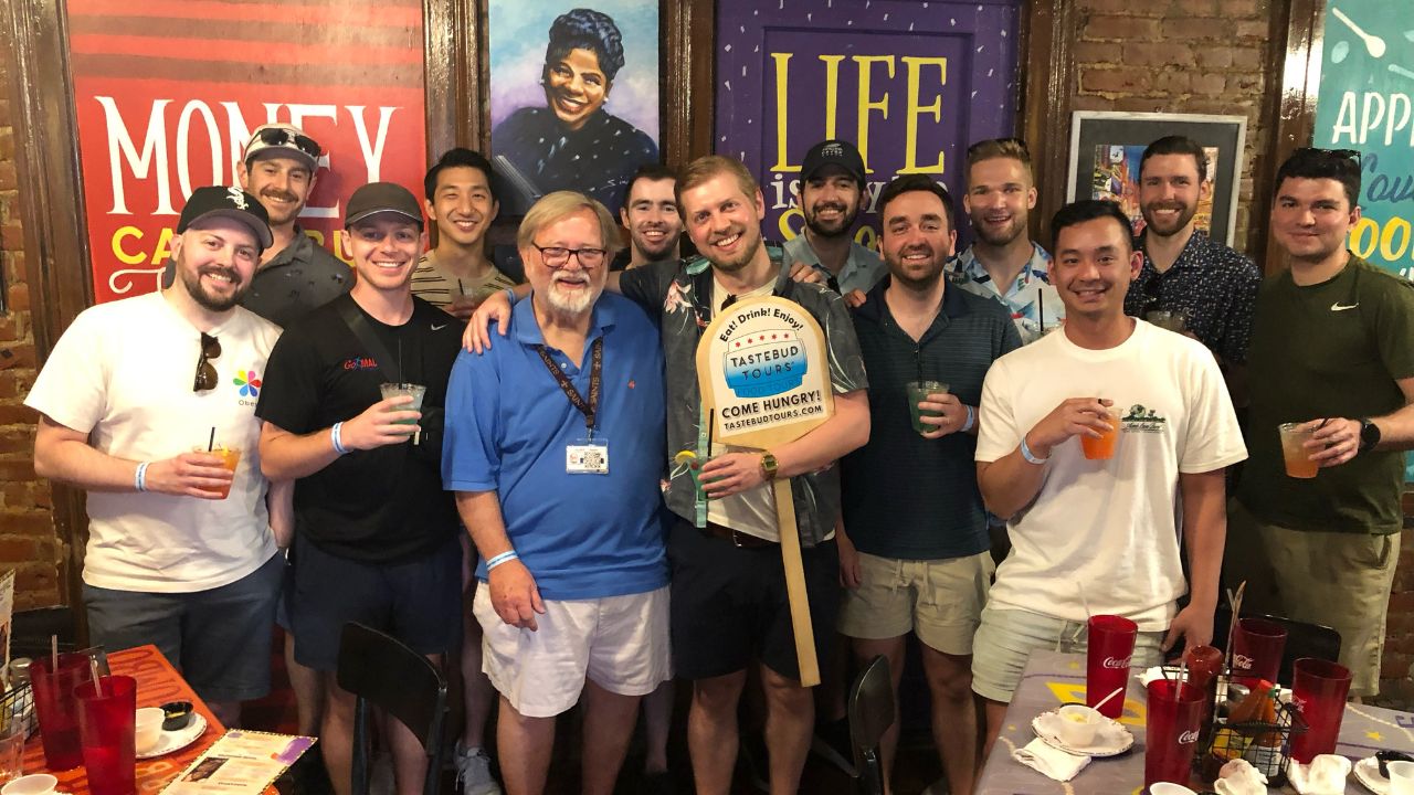 A group of thirteen people pose together indoors, smiling and holding drinks.