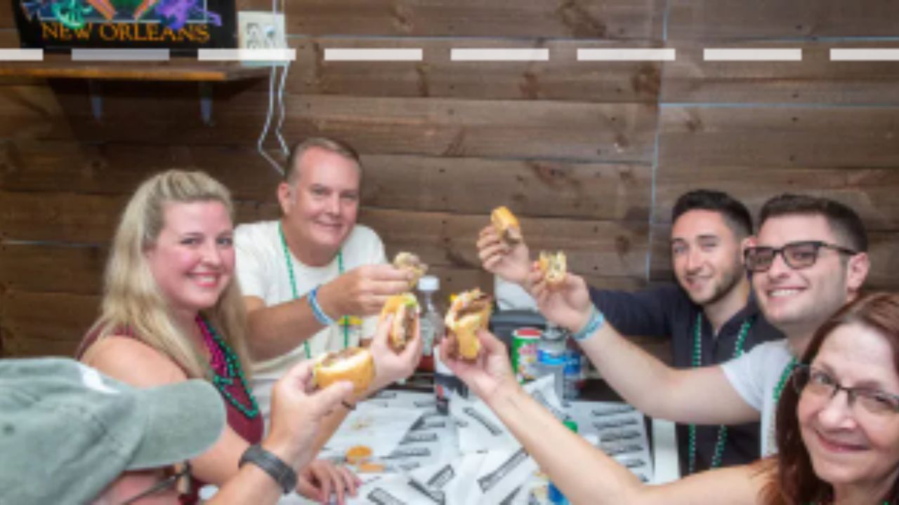 A group of people on a New Orleans food tour raising po-boy sandwiches together in a toast while seated at a rustic wooden table.