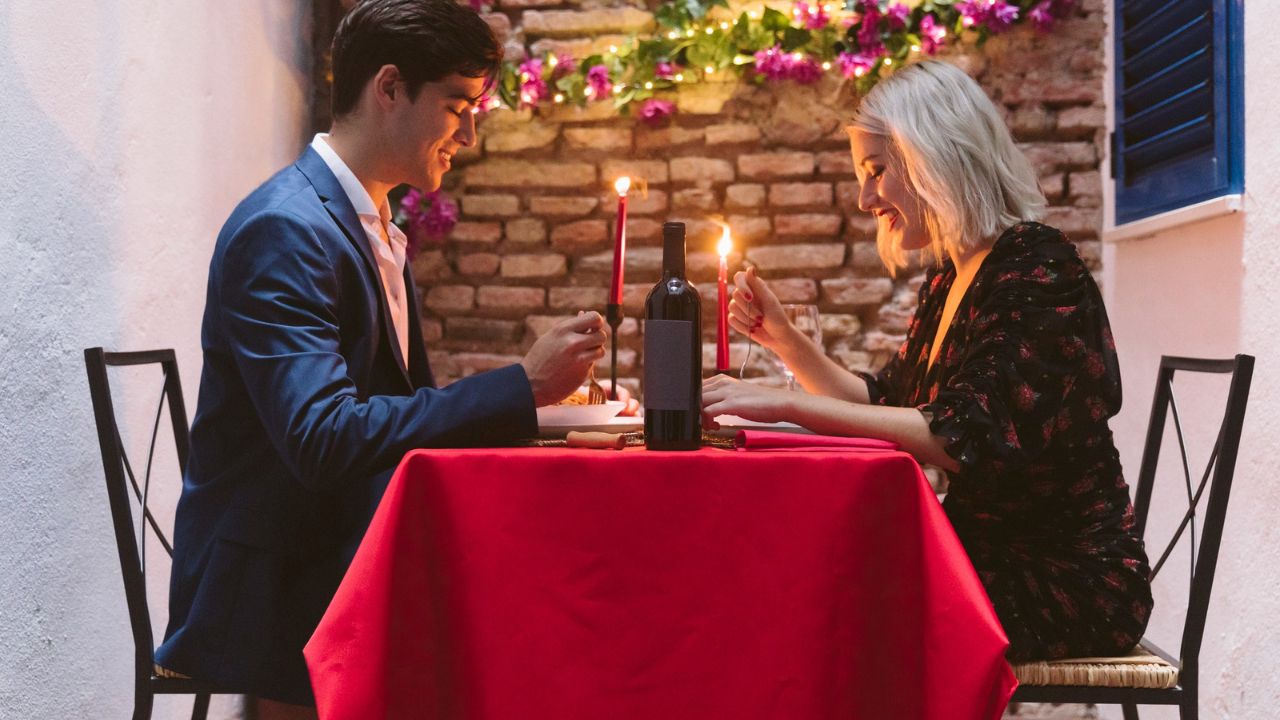 Couple dining at a table with red cloth, candles, and a wine bottle, smiling at each other.