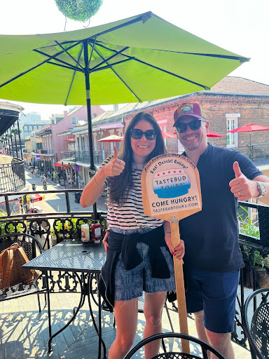 Two people smiling with thumbs up, holding a sign on a balcony with street view.