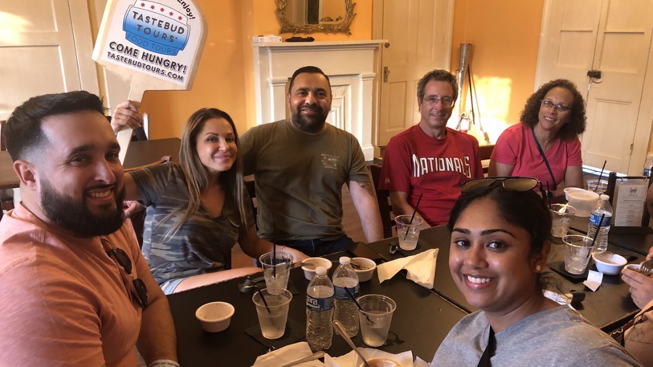 Group of six people at a table, one holding a 'Tastebud Tours' sign.