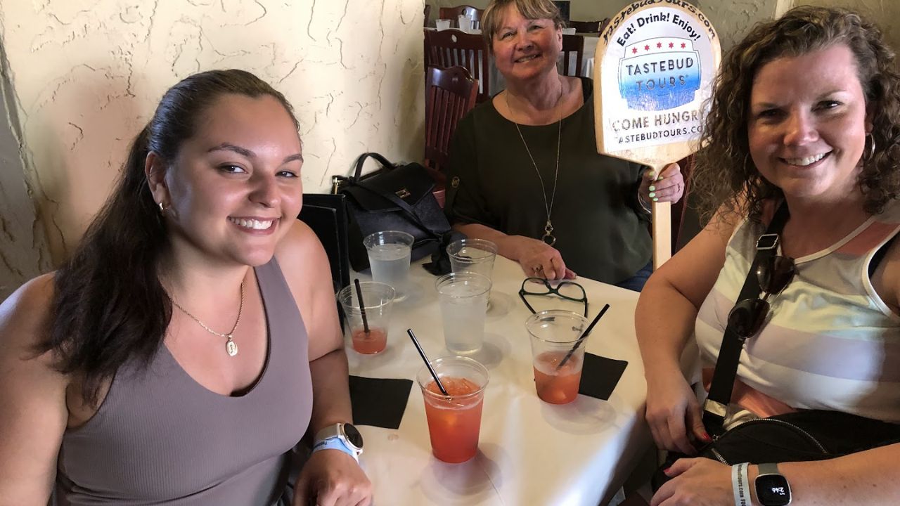 Three women at a table with drinks, one holding a 'Tastebud Tours' sign.