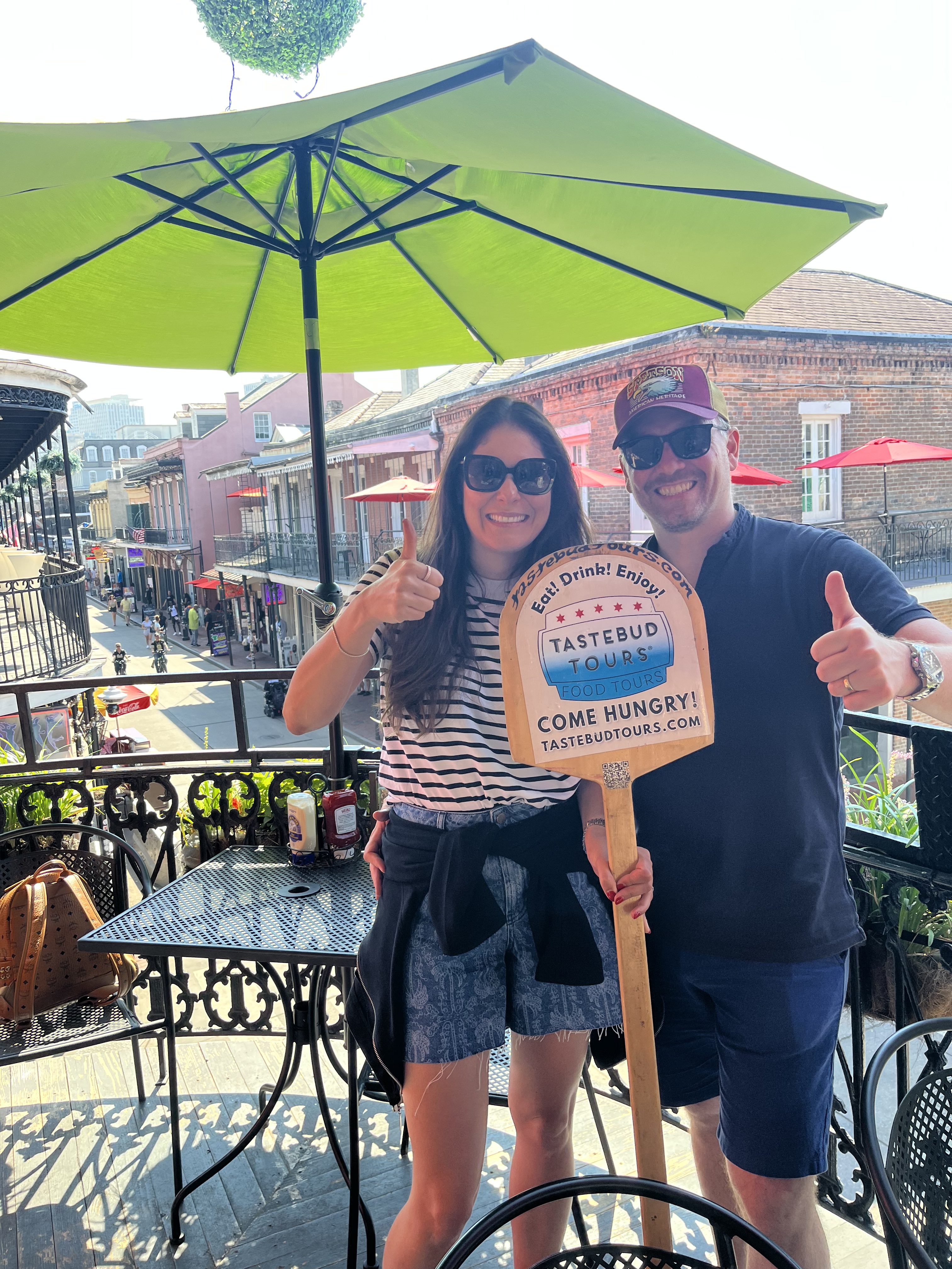 Couple giving thumbs up on a French Quarter balcony with a Tastebud Tours sign.