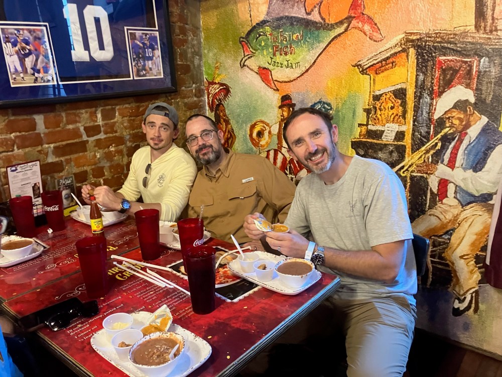 Three men dining at a table with drinks and meals in a restaurant with colorful wall art.