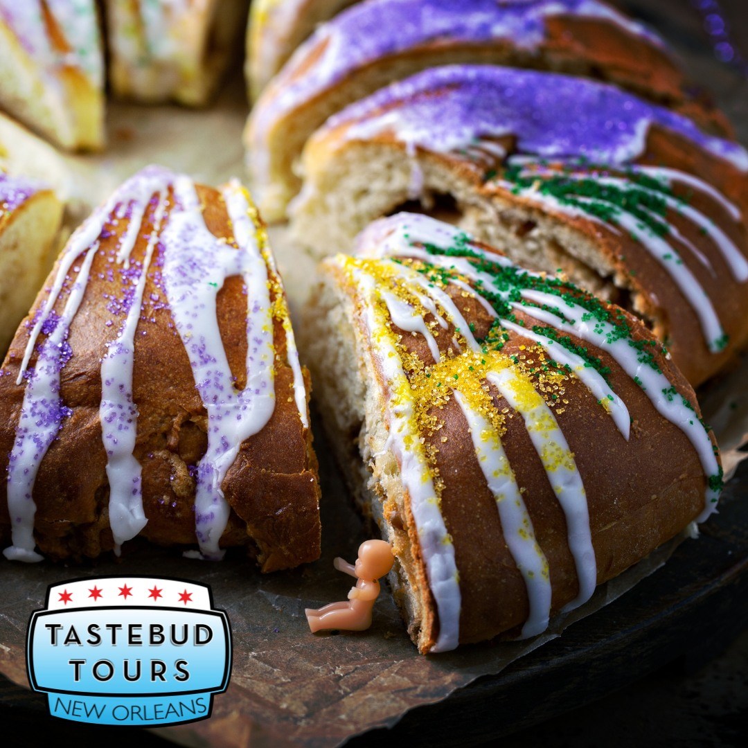Sliced New Orleans king cake topped with white icing and purple, green, and gold sugar, with a small plastic baby beside it.