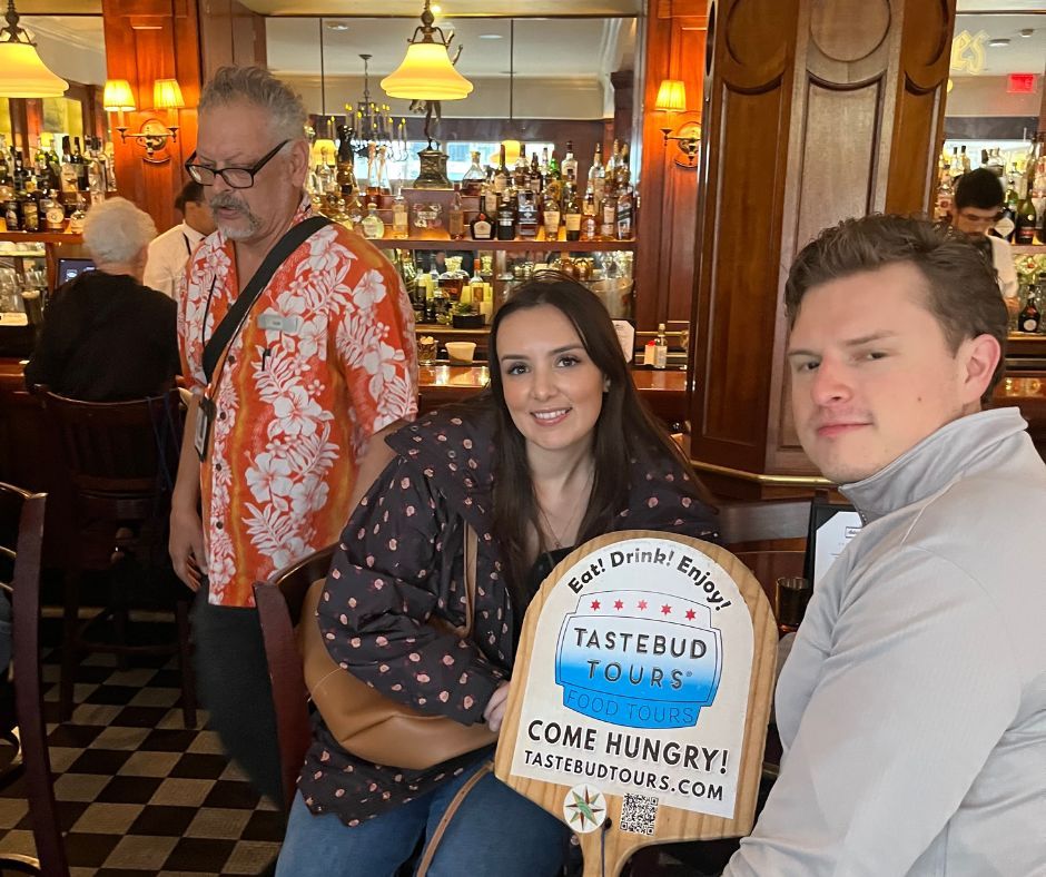 Tastebud Tours guests posing with a food tour sign inside a New Orleans bar with wooden decor and liquor shelves.